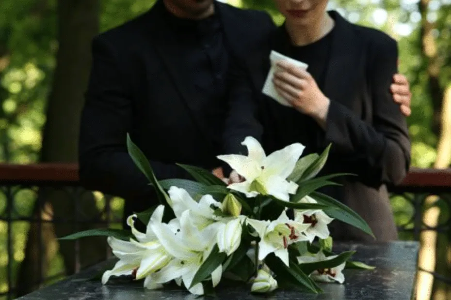 Un uomo in completo nero sostiene emotivamente una donna in abito scuro, entrambi davanti a una bara